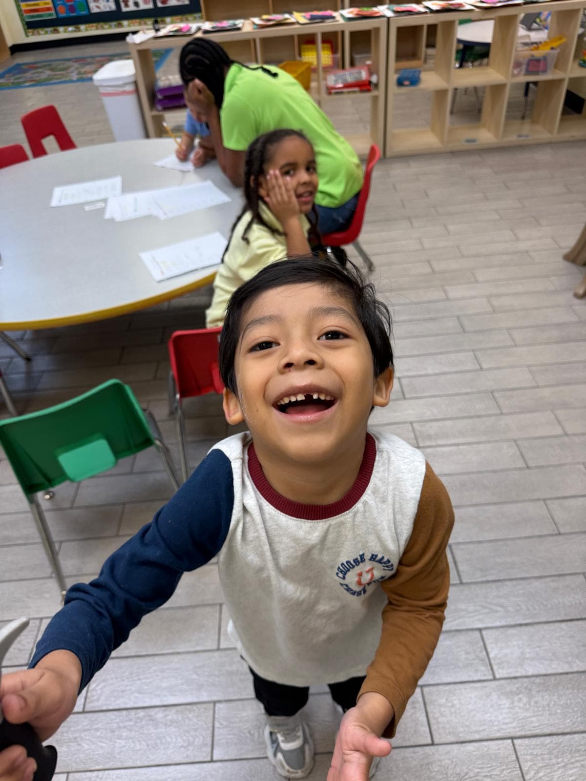 A child smiling in a classroom, with other children and a teacher seated at a table in the background