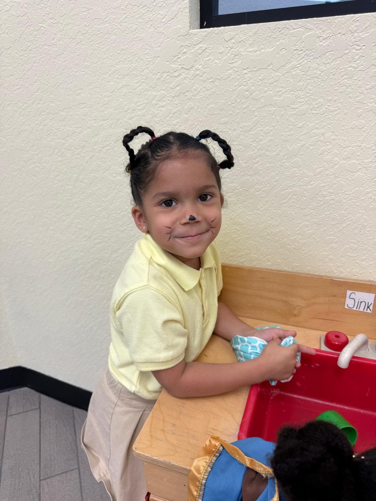 A child at a play kitchen sink with a painted-on whiskers face